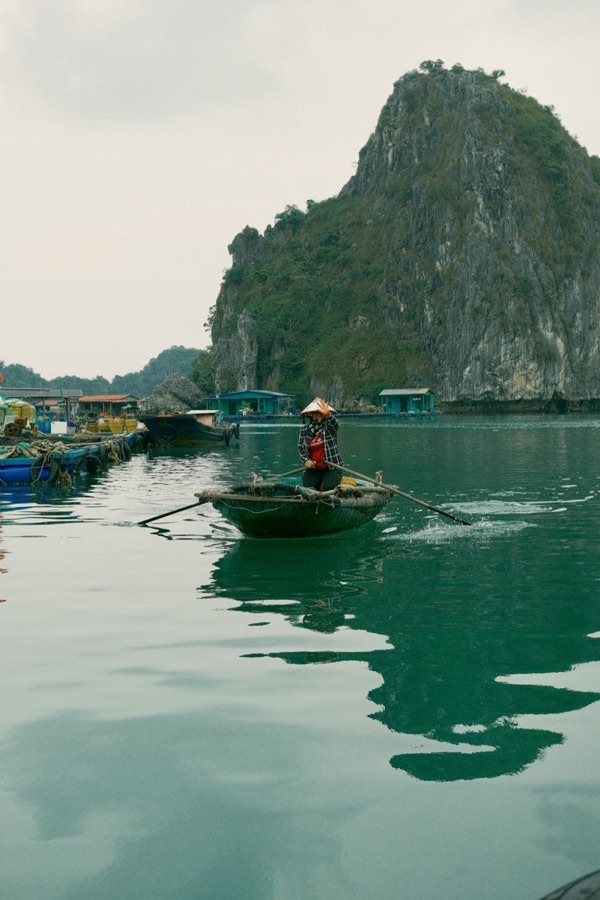 Person rowing a round boat near a floating fishing village with karst limestone peaks in Ha Long Bay, Vietnam