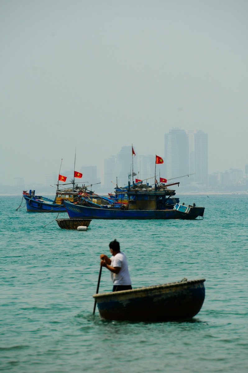 Fisherman rowing a small boat with fishing vessels behind, Hai Phong